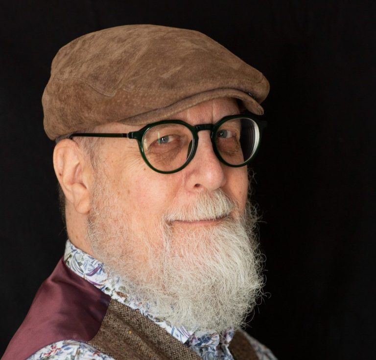 A warm and friendly portrait of a bearded man wearing glasses and a flat cap. He radiates wisdom, experience, and a warm personality