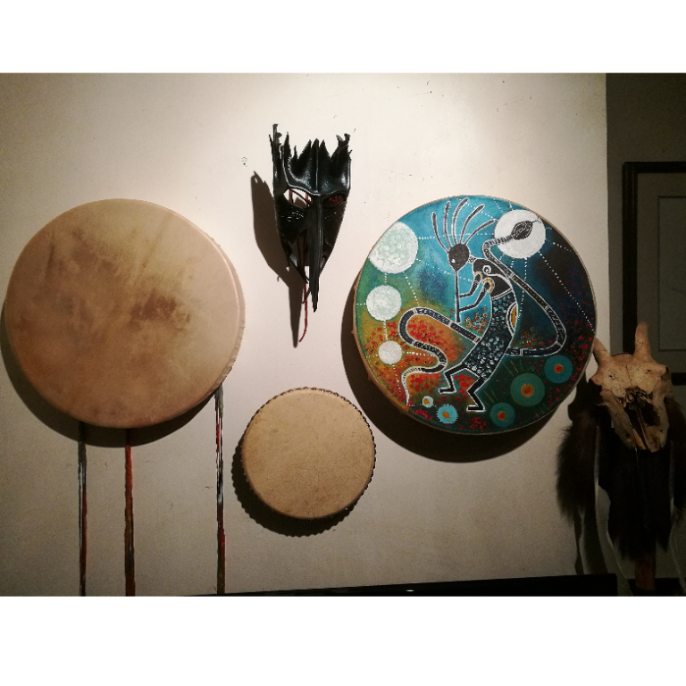 A wall display featuring three ceremonial drums, including a hand-painted shamanic drum with a Kokopelli design, and a leather ritual mask